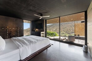 Une chambre à coucher moderne, dotée d’un grand lit, d’un ventilateur de plafond et offrant une vue sur un paysage montagneux grâce à de grandes fenêtres.