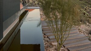 Ein modernes Gebäude mit Wasserspiel und Reflexionen, einem Kiesweg und einer Wüstenlandschaft mit Bergen im Hintergrund.