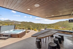 Un patio exterior con jacuzzi, mesa de madera y taburetes, con vista a un paisaje montañoso.