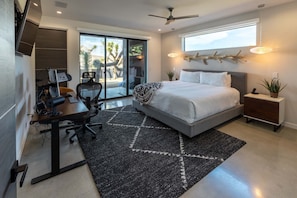 A modern bedroom with a large bed, a desk, and a computer. There is a balcony with a view of palm trees.