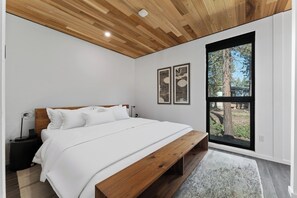 Un dormitorio con techo de madera, un ventanal con vistas a árboles, una cama con ropa blanca y dos cuadros en la pared.