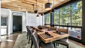 Un espace repas moderne avec une table et des chaises en bois, de grandes fenêtres donnant sur une forêt et une cuisine équipée d’un réfrigérateur.
