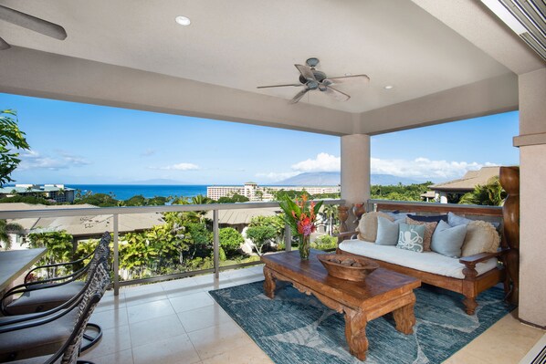 A balcony with a view of the ocean, a ceiling fan, a wooden coffee table, a sofa with cushions, and a vase with flowers. - Waiola Realty - Renovated Tranquil Ho'olei Ocean View Villa 39-2 Wailea Maui (Kihei)