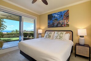 A bedroom with a large bed, two bedside lamps, a painting of a coastal scene, and a view through a glass door to a balcony. - Waiola Realty - Renovated Tranquil Ho'olei Ocean View Villa 39-2 Wailea Maui (Kihei)