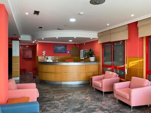A hotel reception area with a curved reception desk, a reception sign, a fire extinguisher, a potted plant, and two pink armchairs. - Hotel Acquamaris 3000 (Sanxenxo)