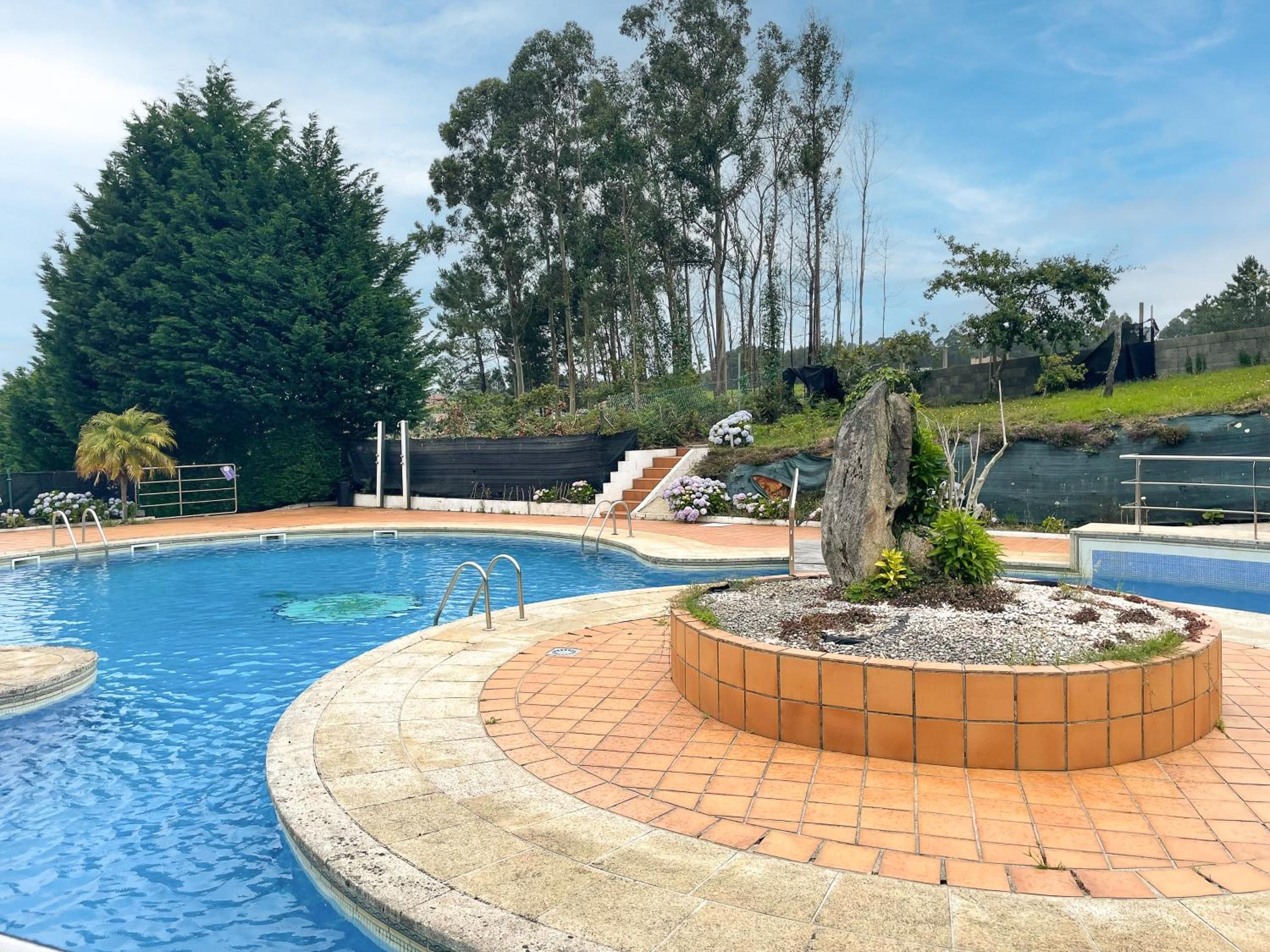 Piscina con borde de baldosas, rodeada de árboles y un jardín ajardinado con una formación rocosa.