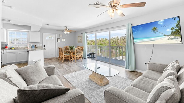 A spacious living room with a glass coffee table, grey sofas, and a balcony with a view of the beach. - Huge Shared Pool, Walk to Beach, Times Square Restaurants! (Fort Myers Beach)