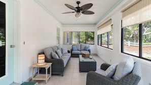 A sunlit room with a ceiling fan, wicker furniture, and large windows offering a view of the outdoors. - Spacious Dog Friendly Home w/ Full Sized Pool - Game Room In Garage- 3BR, 2BTH, (Oak Island)