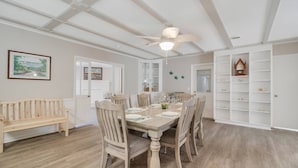 A dining area with a wooden table set for four, surrounded by wooden chairs. A ceiling fan is visible above the table. - Spacious Dog Friendly Home w/ Full Sized Pool - Game Room In Garage- 3BR, 2BTH, (Oak Island)