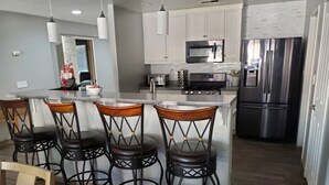 A modern kitchen with white cabinets, a stainless steel refrigerator, a microwave, and a built-in oven. There are four bar stools with wooden seats and metal frames.