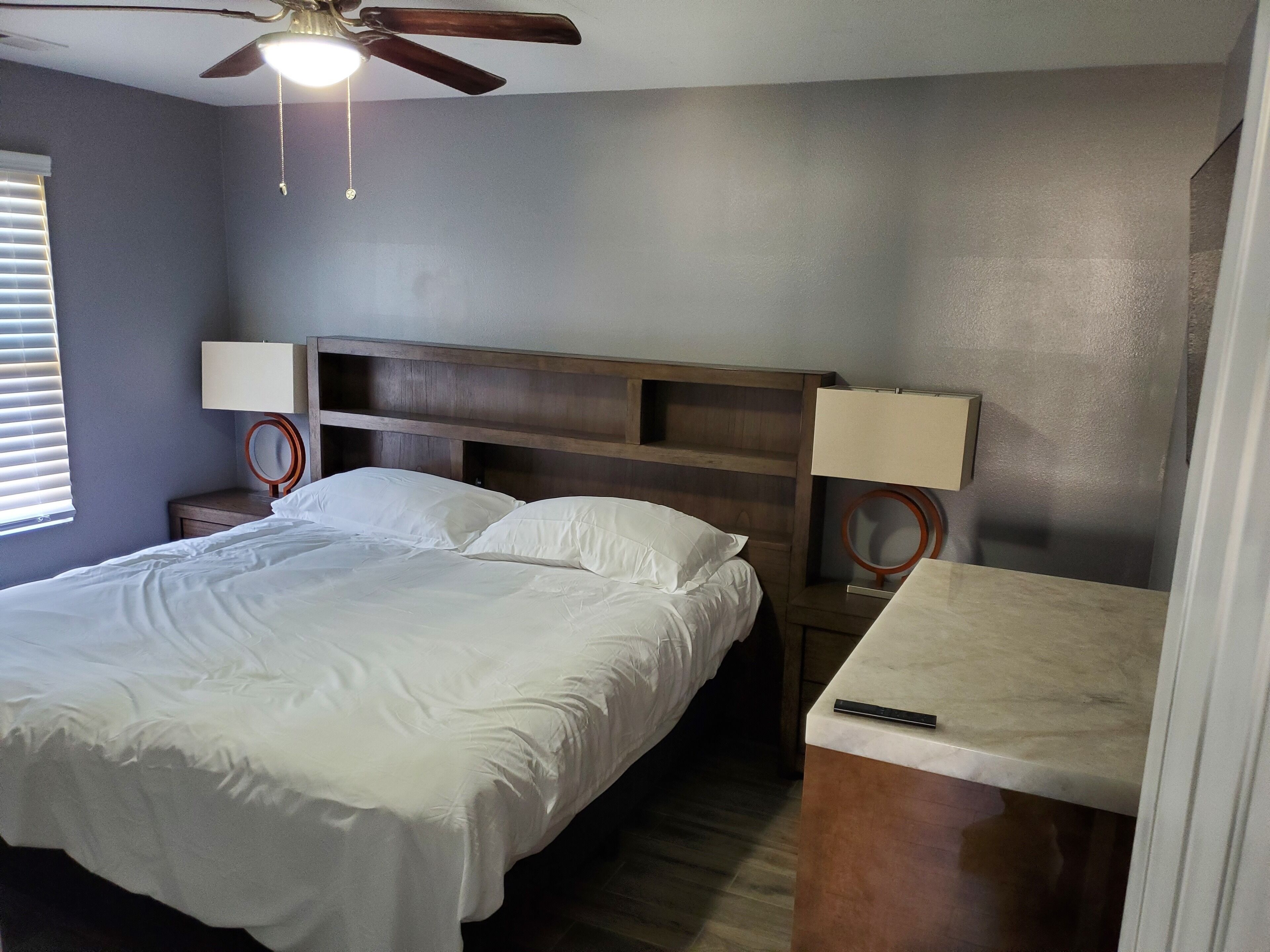 A bedroom with a bed, a nightstand, a desk, a ceiling fan, and a wall-mounted lamp.