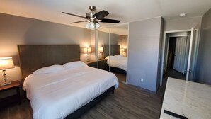 A hotel room with a large bed, a ceiling fan, a nightstand with a lamp, a mirror, and a door leading to another room.