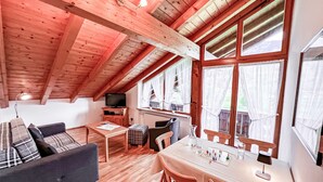 Een knus vertrek met een houten plafond, een bank, een eettafel met stoelen en grote ramen voorzien van gordijnen.