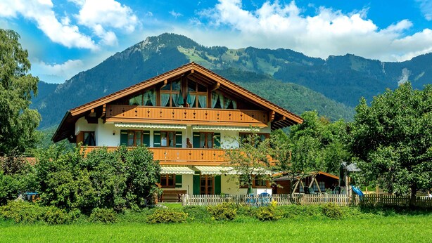Een houten chalet met twee verdiepingen en balkons, omgeven door weelderig groen en bergen op de achtergrond.