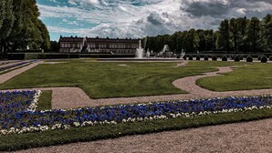 Een formele tuin met een fontein, strak bijgesnoeide hagen en een groot gebouw op de achtergrond.