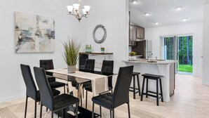 A modern kitchen with a central island, a dining area with a chandelier, and a view of a lush garden through a sliding door.