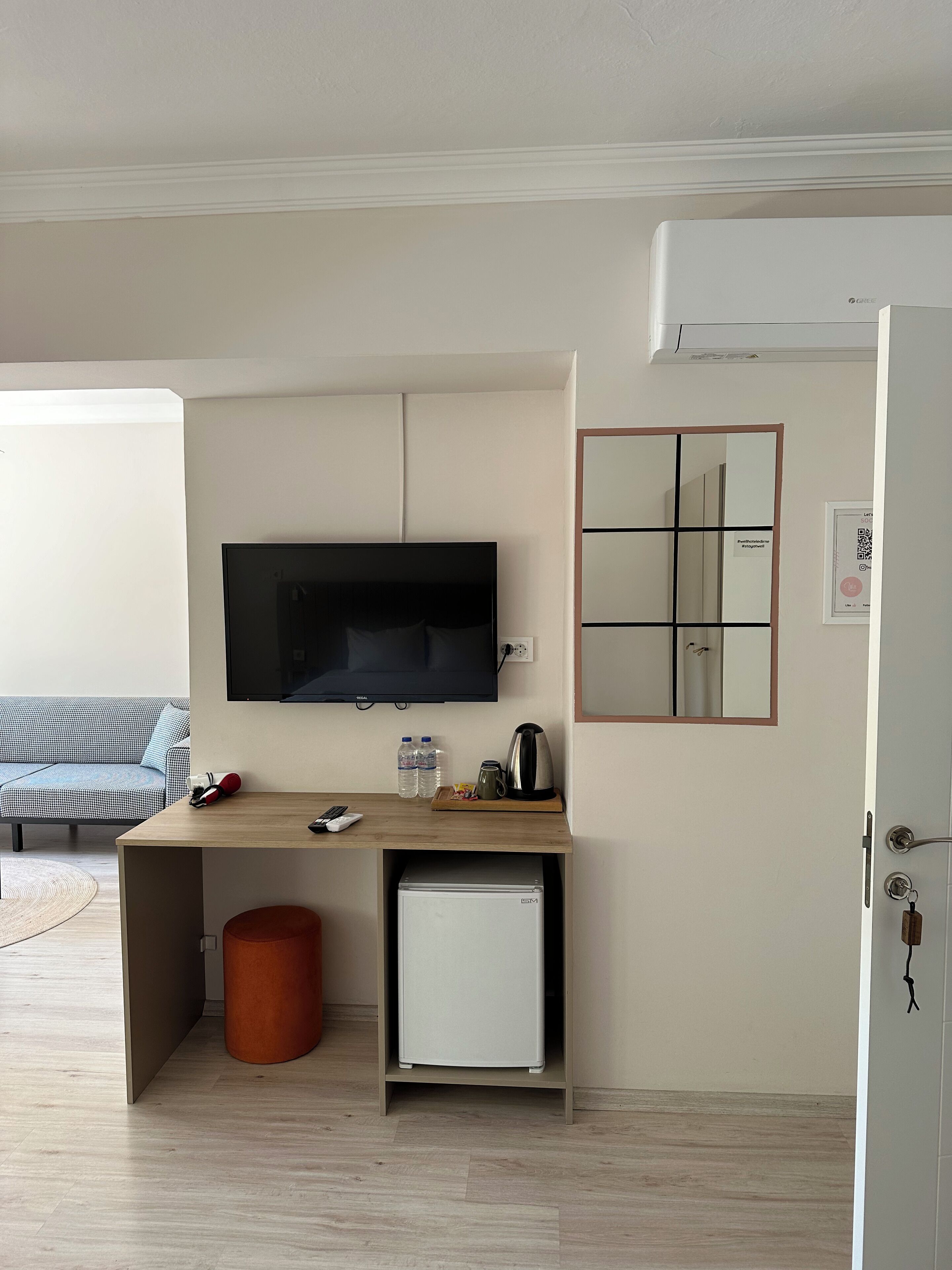 A small, modern room with a TV mounted on the wall, a small fridge, a wooden desk with a kettle, and a mirror.