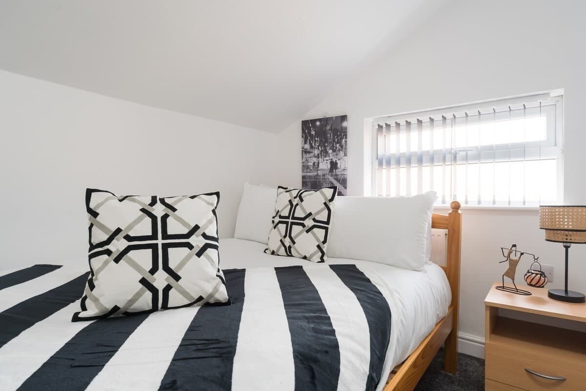 Un dormitorio con cama, almohadas, una mesita de noche, lámpara y ventana con persianas.