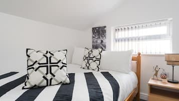 Un dormitorio con cama, almohadas, una mesita de noche, lámpara y ventana con persianas.