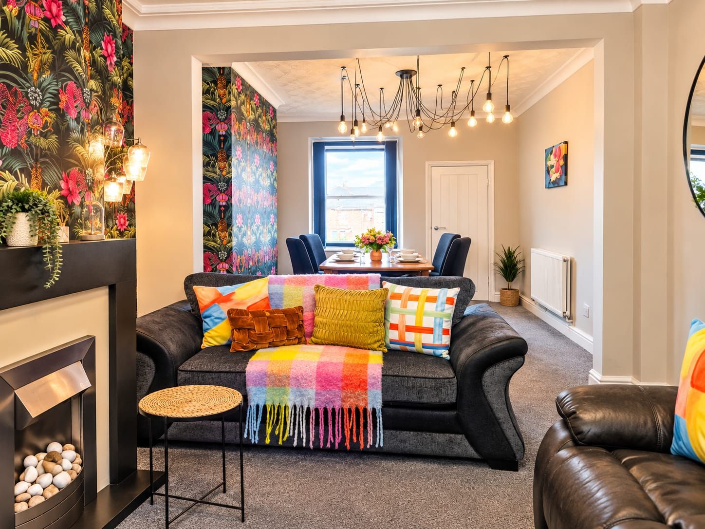 A cozy living room with a fireplace, a sofa with colorful cushions, a dining area with chairs, and a large window providing natural light.
