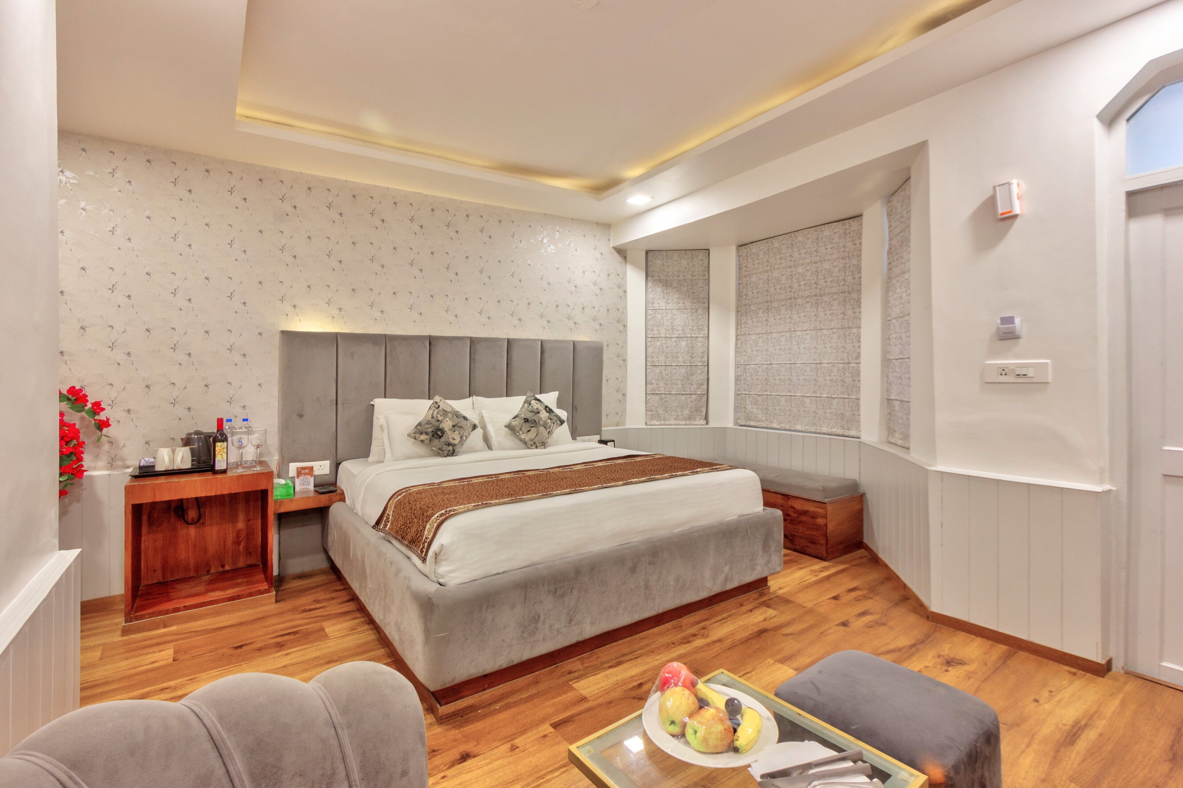 A modern bedroom with a large bed, bedside tables, a small seating area, and a window with blinds.