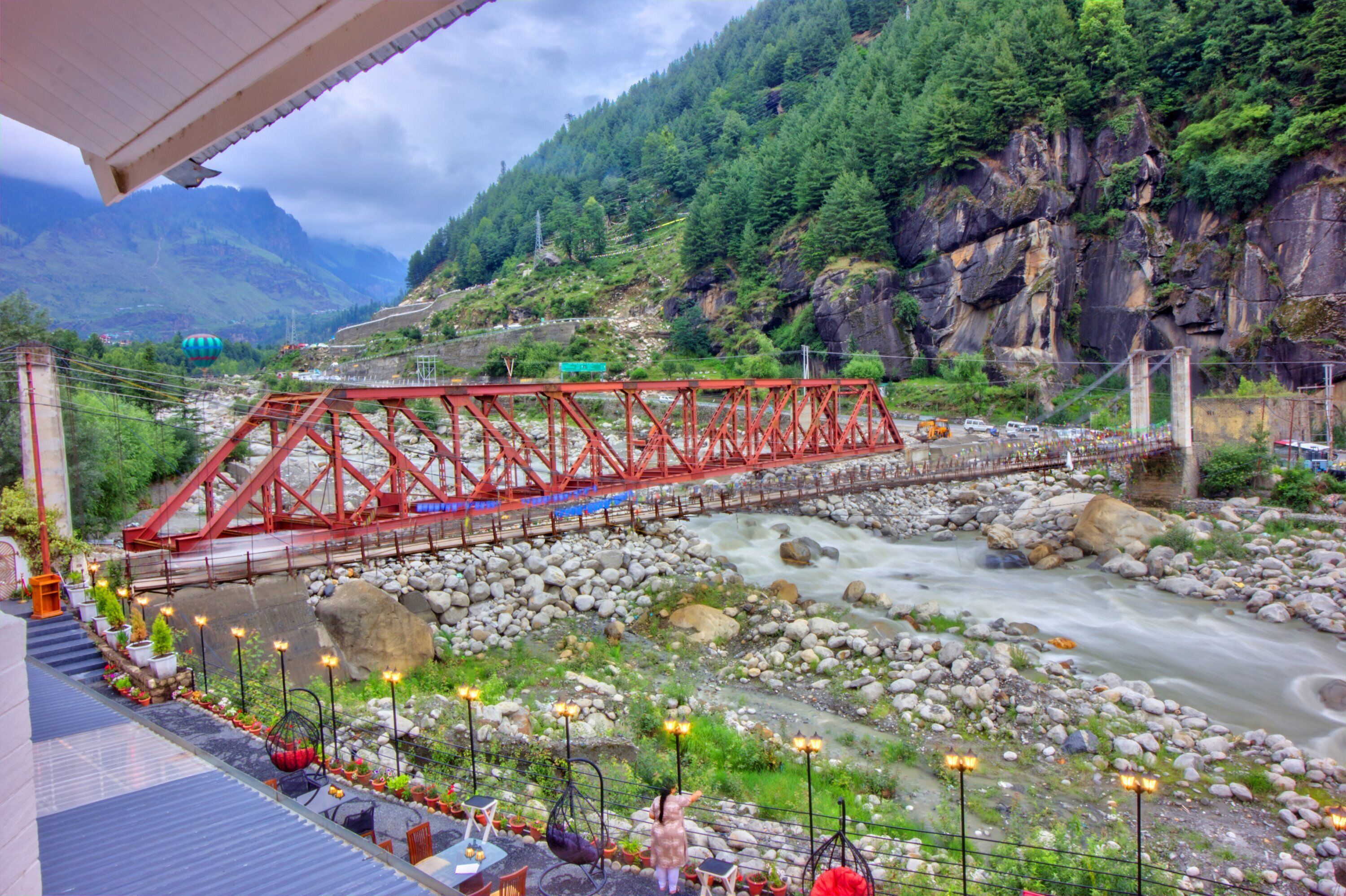 Un puente rojo sobre un río con lecho pedregoso, rodeado de vegetación y montañas.