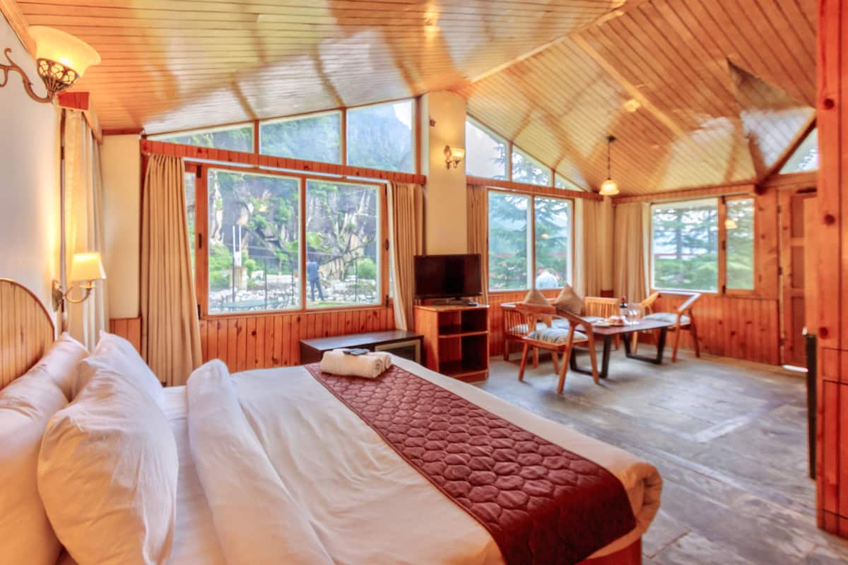 A spacious room with a large bed, a desk, and a television. There are large windows providing a view of the outdoors.