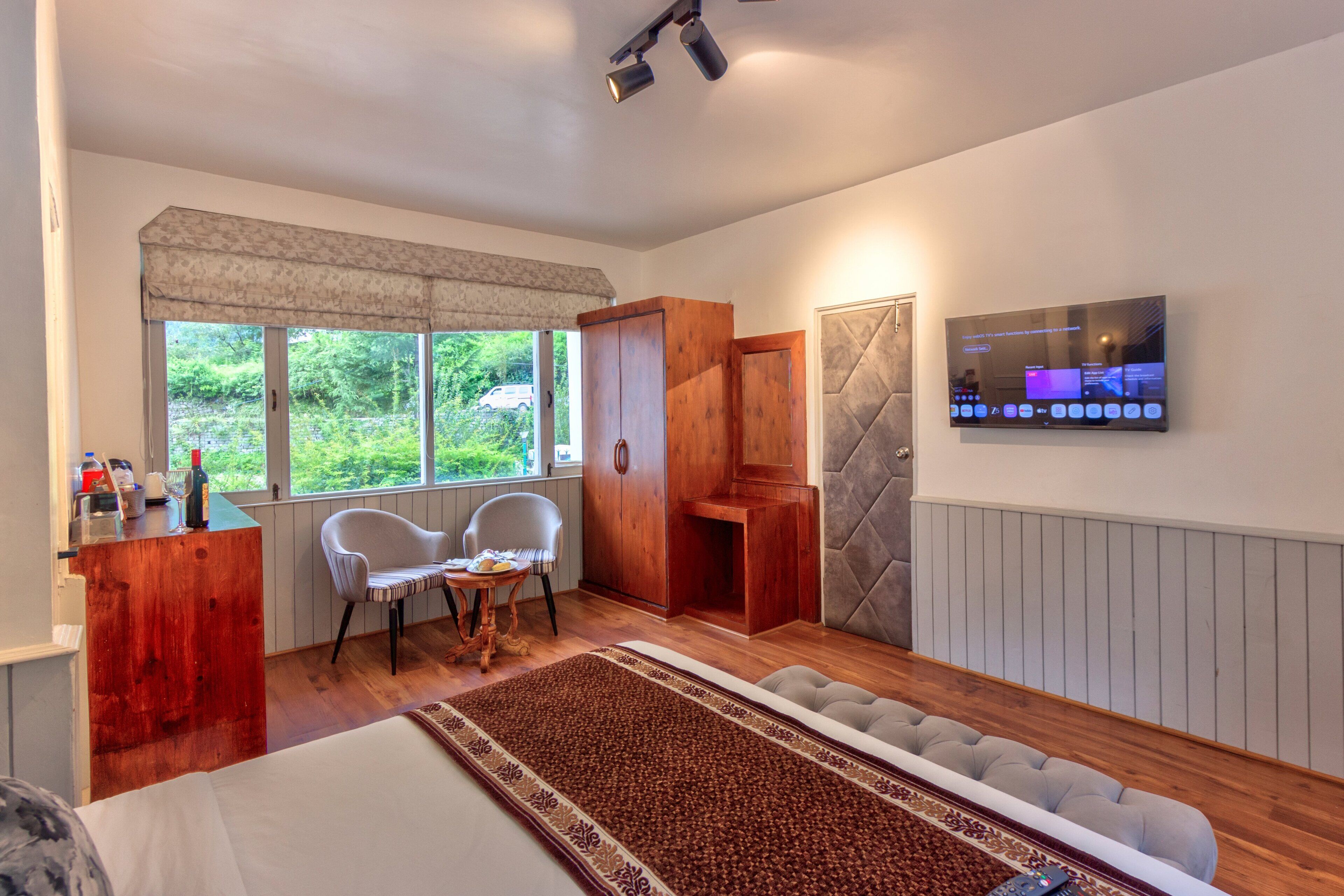 A bedroom with a bed, a desk, a chair, a TV, and a window with a view of greenery.