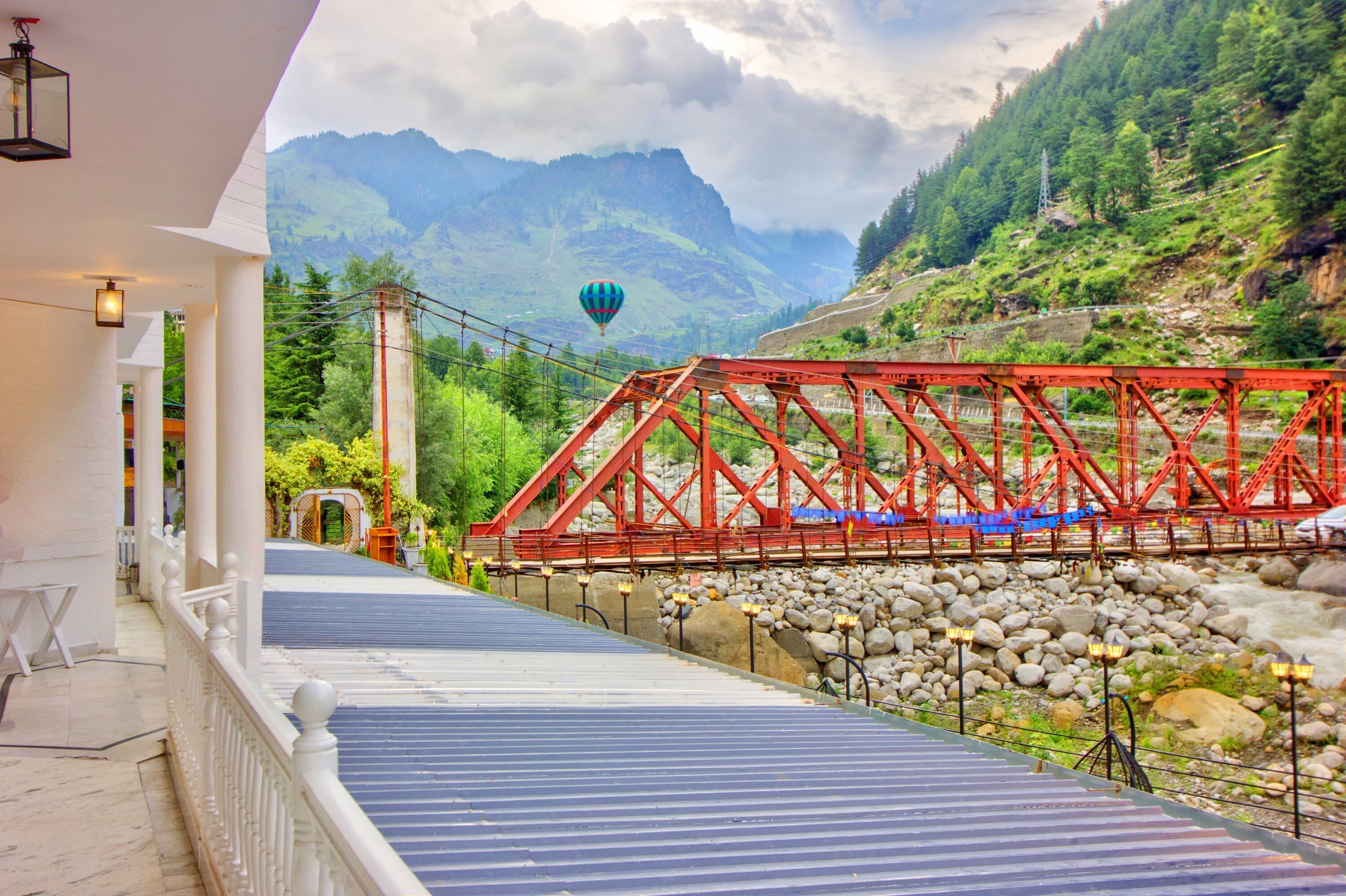 A red bridge over a river, with a hot air balloon in the background and a building with a balcony in the foreground.