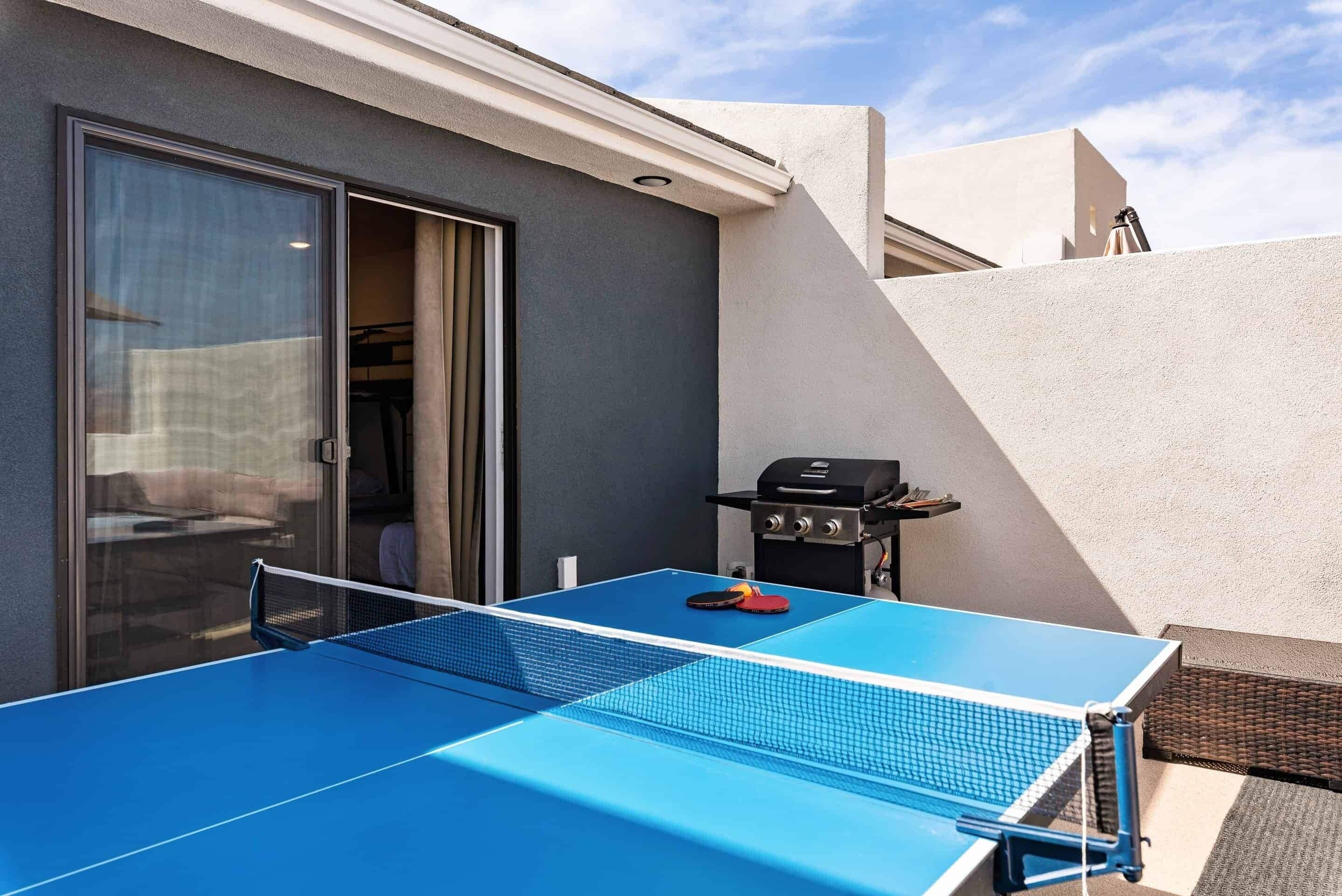 A blue ping pong table with a net, a grill, and a sliding glass door.