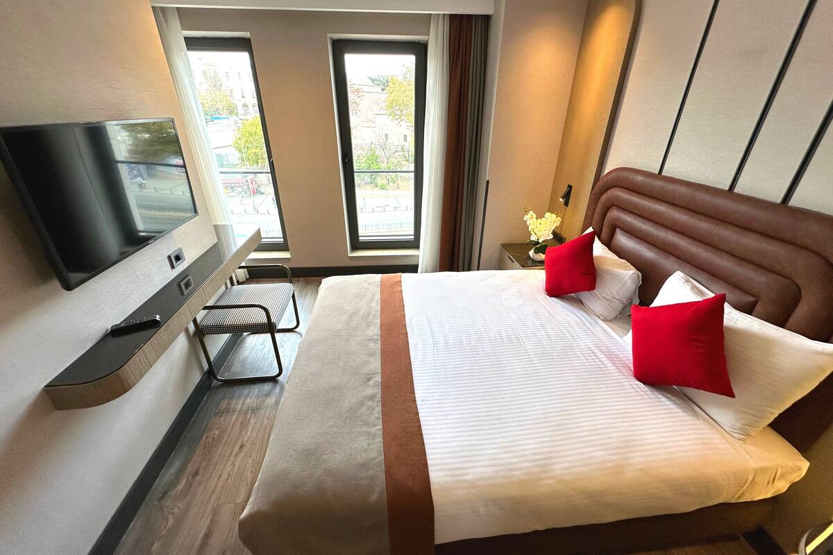 Une chambre d’hôtel équipée d’un lit, d’une télévision, d’un bureau et d’une fenêtre donnant sur des immeubles.