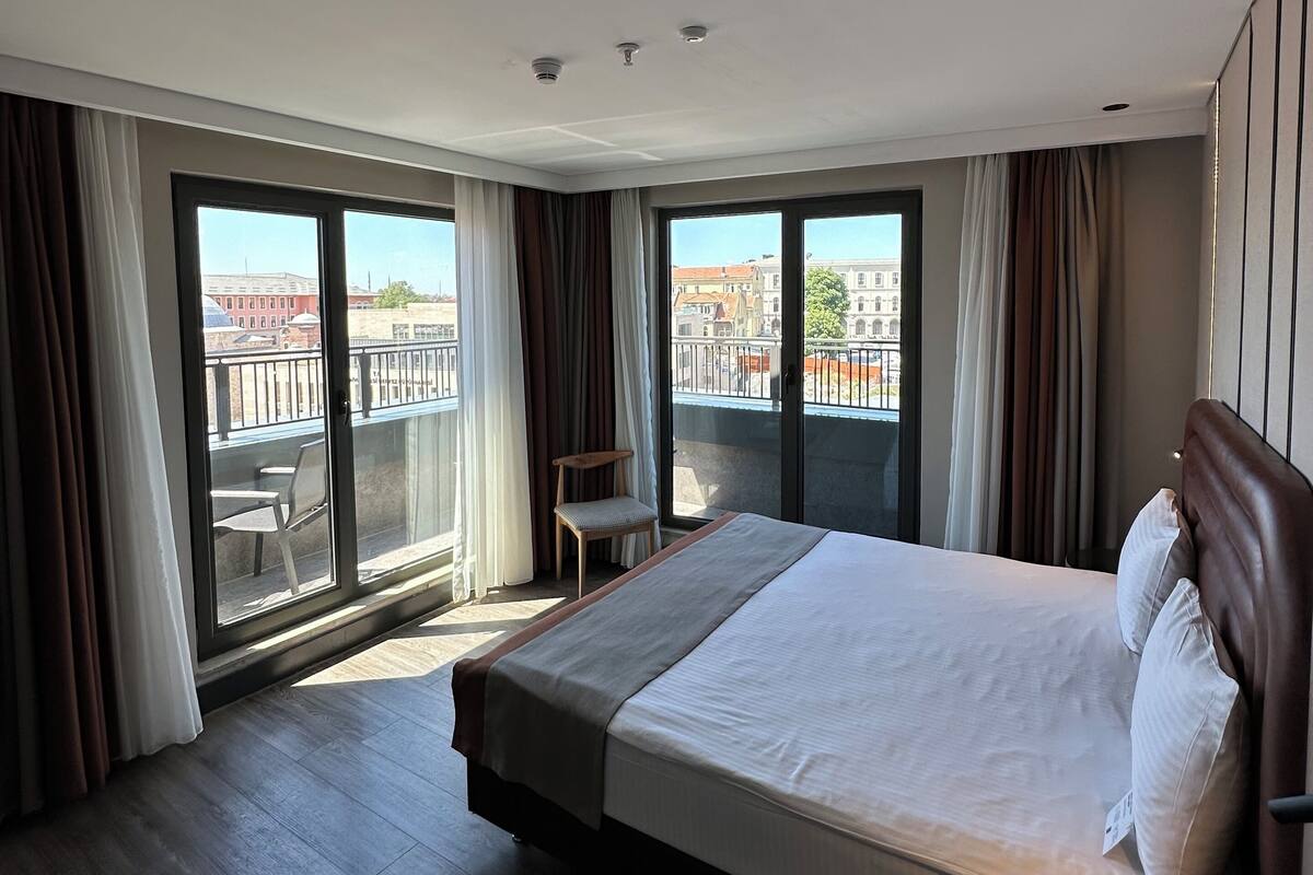 Une chambre d’hôtel moderne avec un grand lit, un petit bureau et un balcon donnant sur les immeubles.