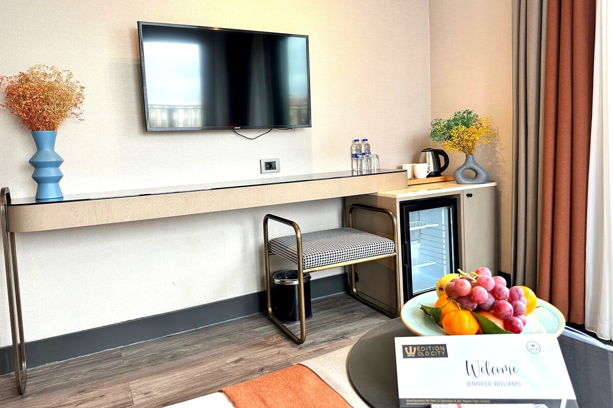 Une chambre d’hôtel moderne équipée d’un téléviseur à écran plat fixé au mur, d’un petit bureau avec une chaise, d’un mini-réfrigérateur et d’une pancarte de bienvenue.