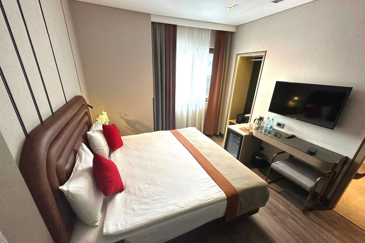 Une chambre d’hôtel comprenant un lit, un bureau avec un téléviseur à écran plat, une petite kitchenette et une fenêtre avec des rideaux.