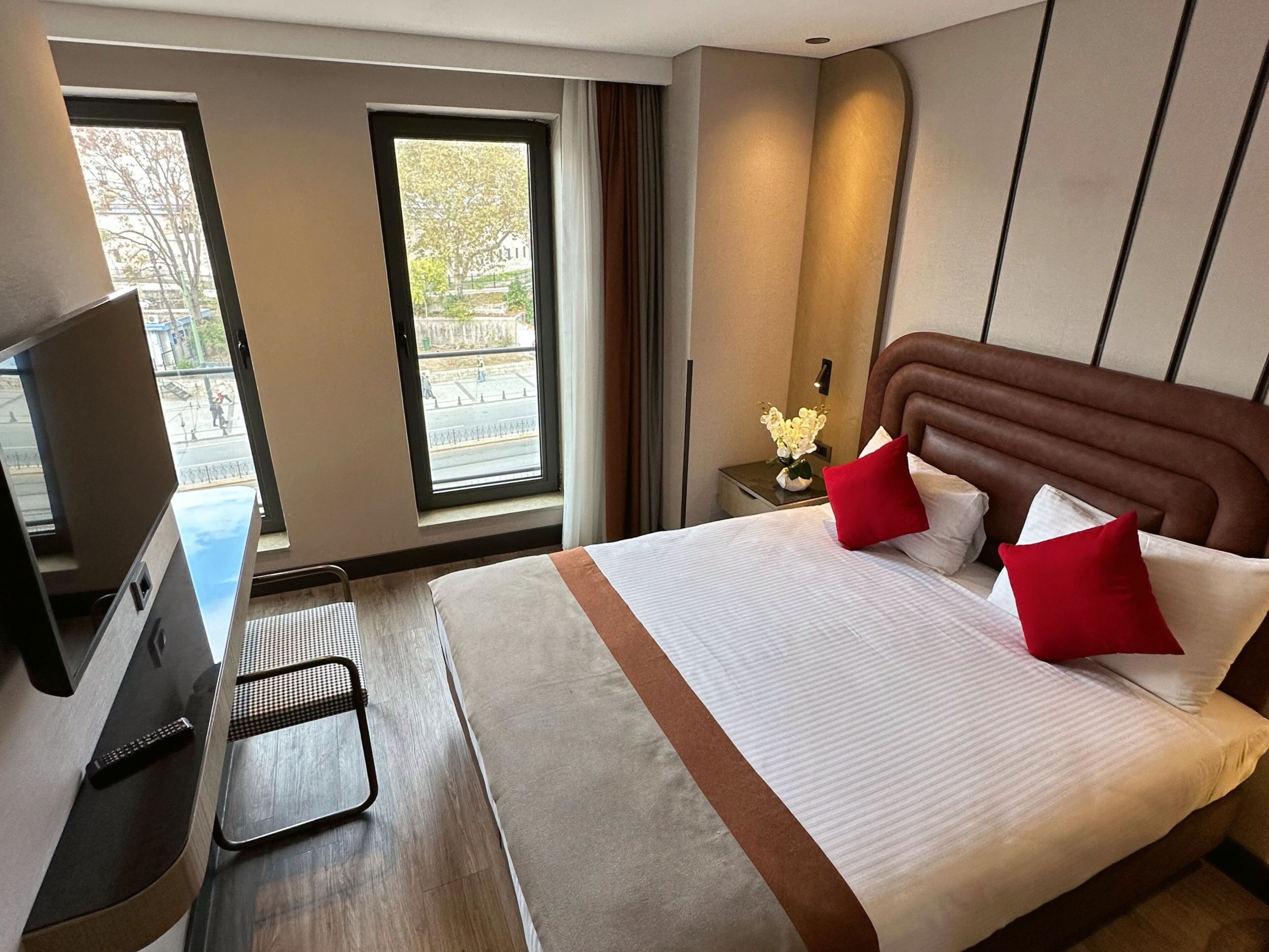 A hotel room with a bed, a TV, a desk, and a window with a view of the street.