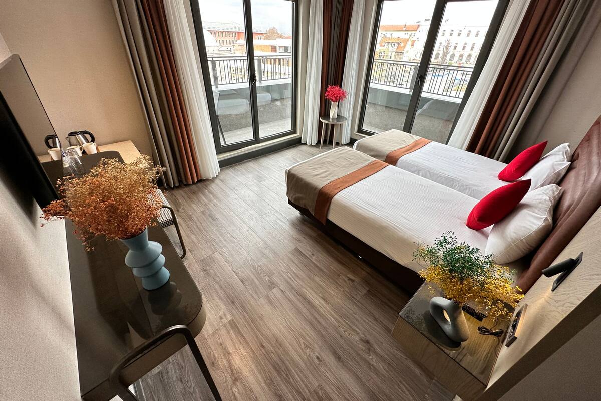 Une chambre d’hôtel avec un grand lit, un bureau avec une chaise, une petite table ornée d’un vase de fleurs et une vue sur la ville grâce à de grandes fenêtres.