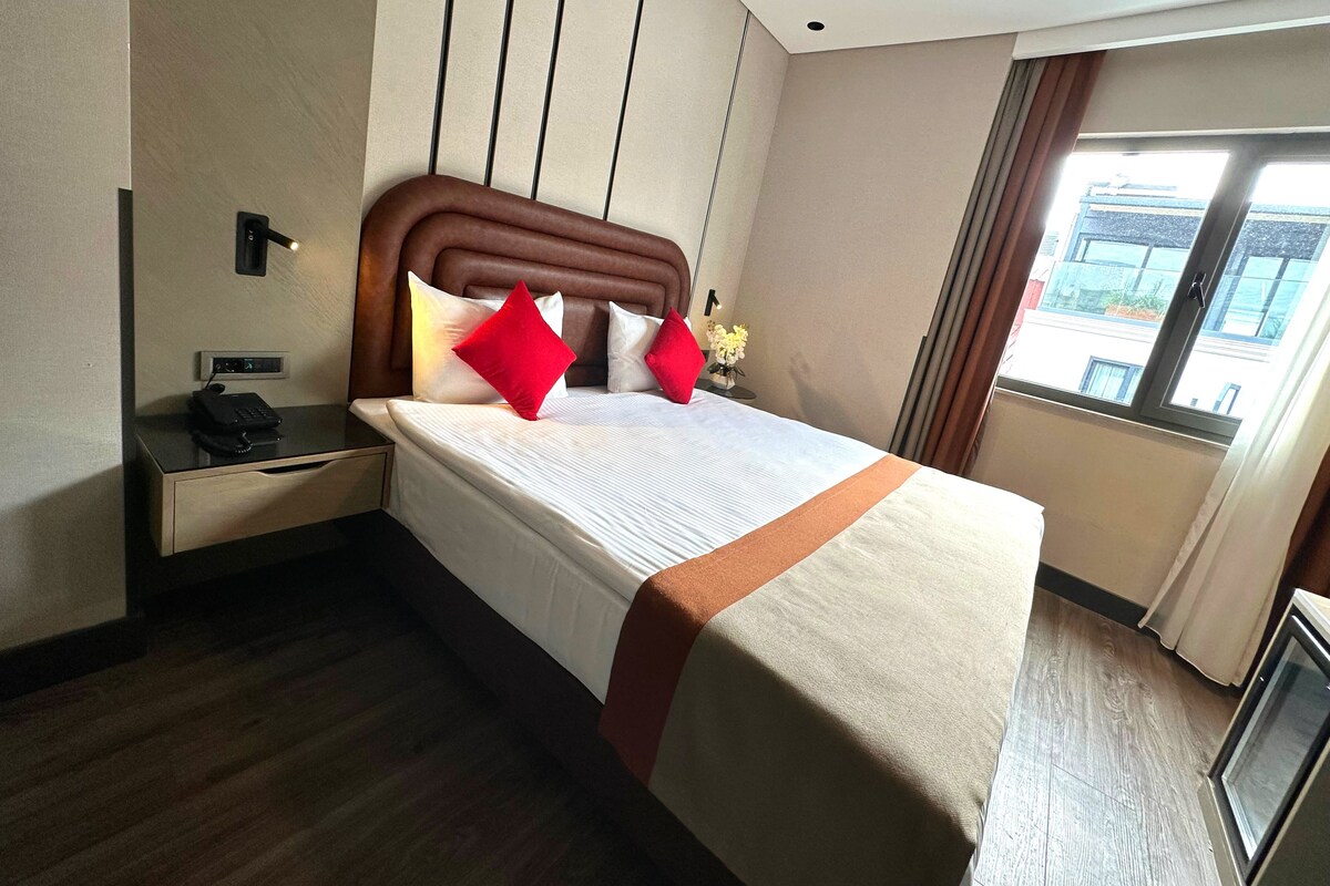 Une chambre d’hôtel avec un grand lit, une table de chevet avec un téléphone, une commode et une fenêtre donnant sur des immeubles.