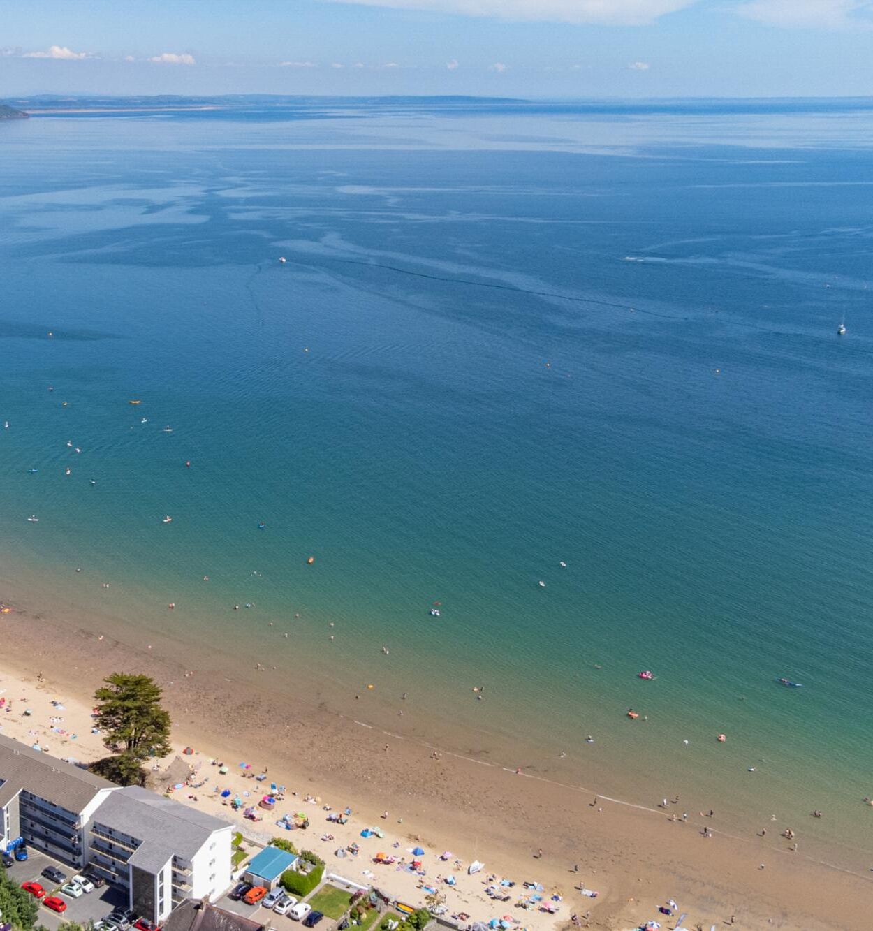 Vista aèria d'una platja amb aigües cristal·lines, sorra daurada i un edifici de diverses plantes amb pàrquing.