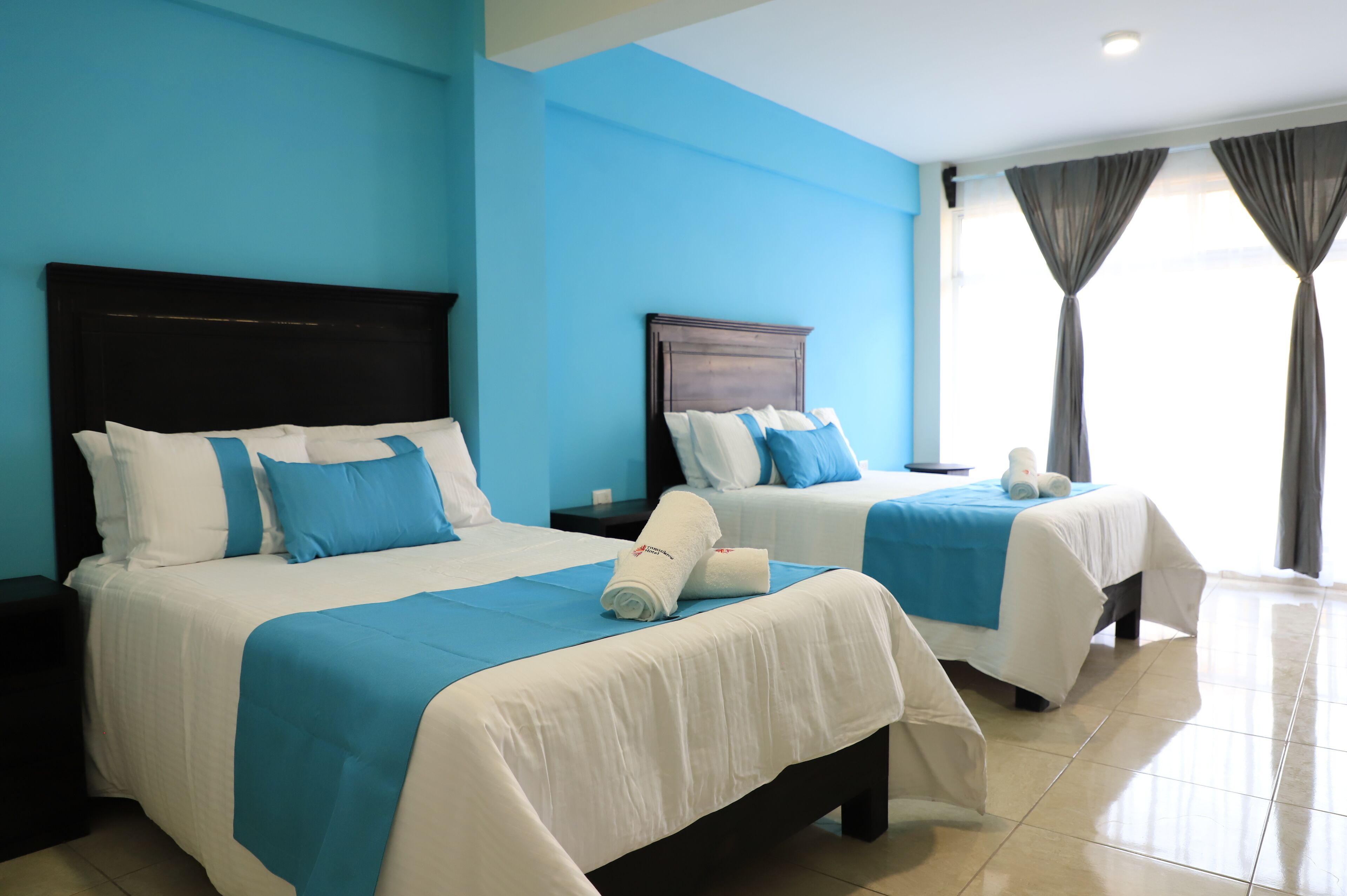 Dos camas individuales con ropa de cama azul y blanca, cabecera oscura y una ventana con cortinas.