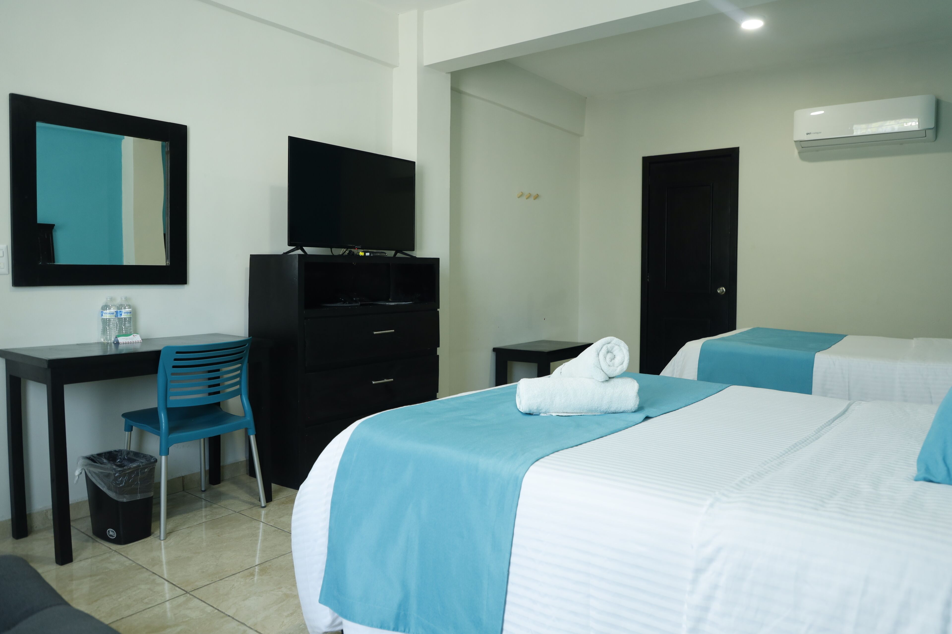 A hotel room with a bed, a desk, a chair, a TV, a trash bin, and an air conditioning unit.