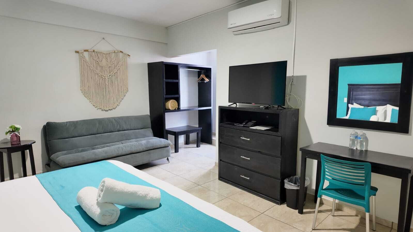 Una habitación de hotel moderna con un sofá gris, una cama con cubrecama azul, un escritorio con silla, un armario y un televisor en la pared.