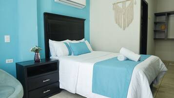 Una cama bien tendida con colcha blanca y azul, una mesita de noche negra con cajones y un aire acondicionado instalado en la pared.