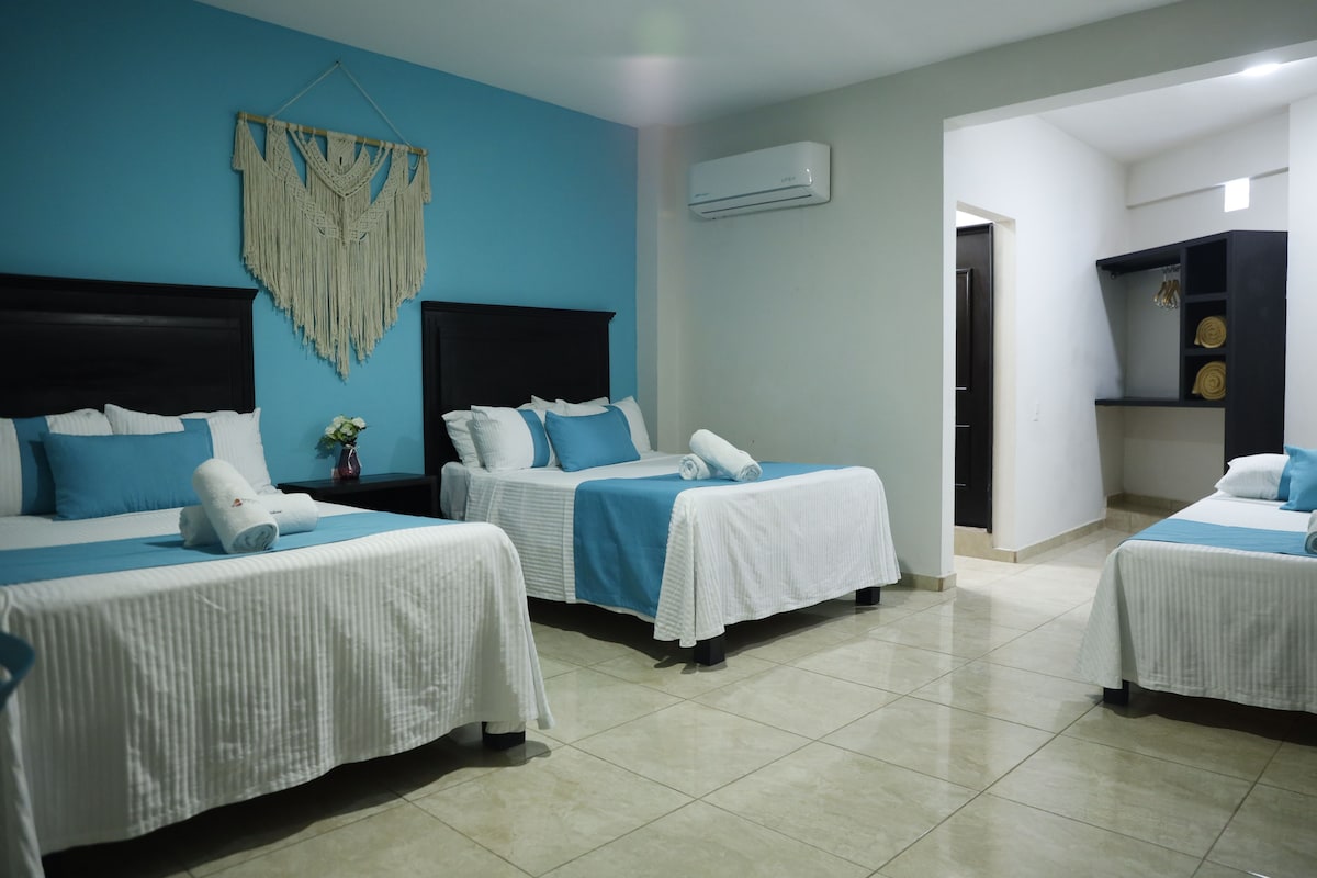 A hotel room with three beds, a macrame wall hanging, an air conditioning unit, and a hallway leading to a bathroom.