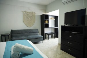 Habitación de hotel con cama, sofá gris, televisor, armario, pequeña mesa y un tapiz de macramé en la pared.