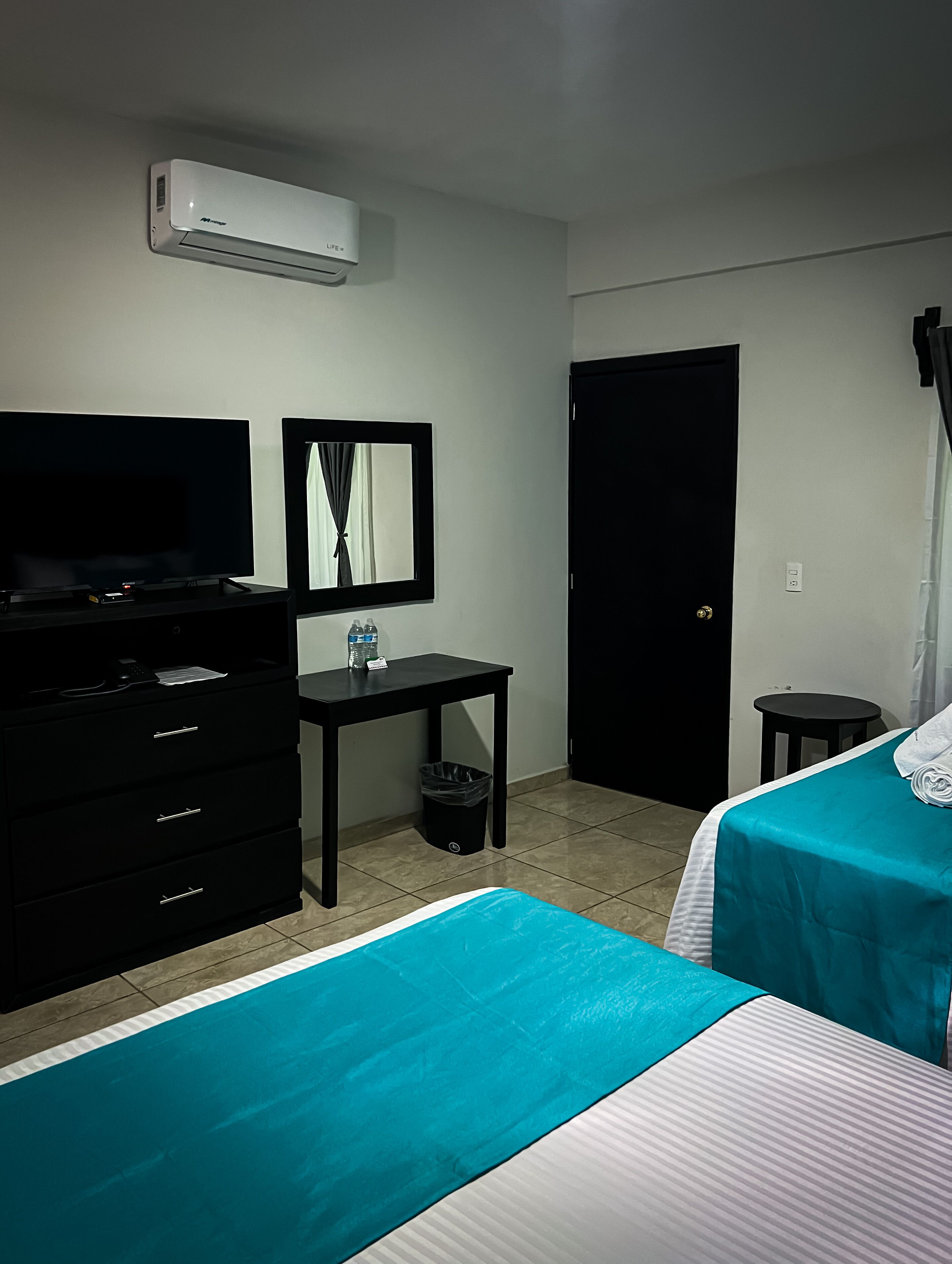 Una habitación de hotel con una cama, televisión, escritorio, cubo de basura y una puerta.