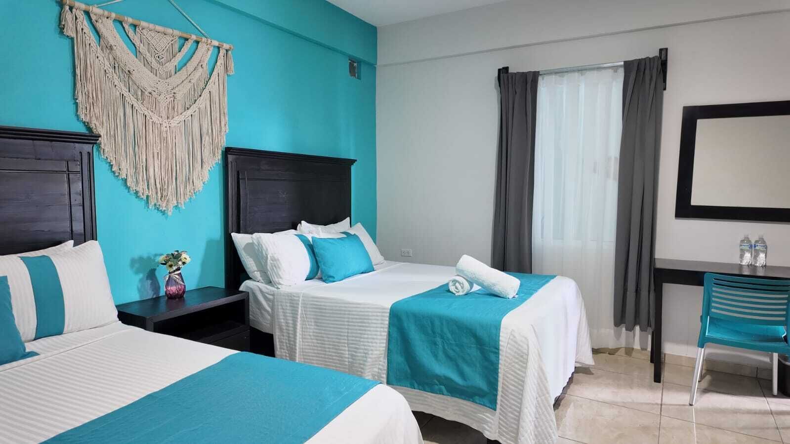 A hotel room with two beds, a desk, a chair, a macrame wall hanging, and a large mirror.