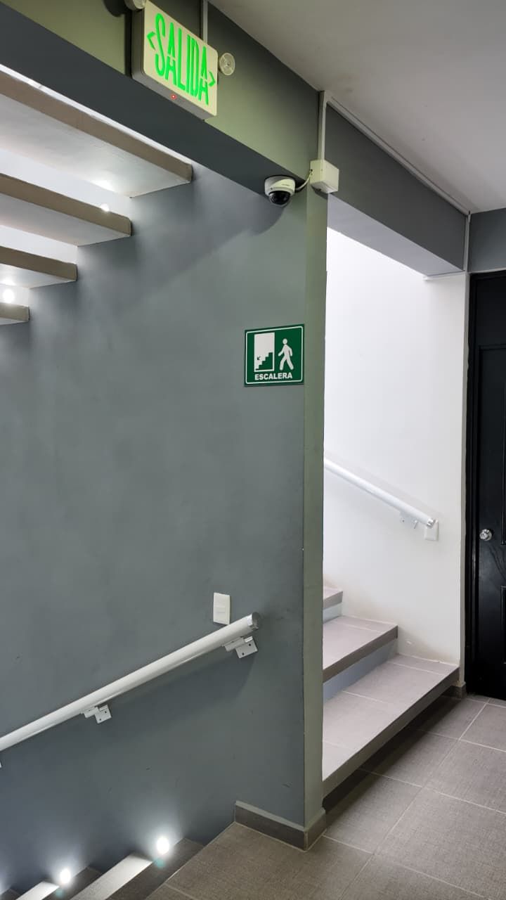 A modern stairwell with a green emergency exit sign, a handrail, and a security camera.