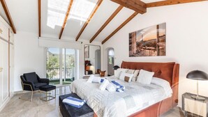 Une chambre à coucher avec un grand lit, une chaise, un banc, une table de chevet et une vue sur l’extérieur.