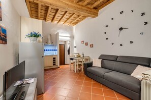 Un salon avec un canapé, un coin repas avec une table et des chaises, une cuisine avec des placards, une horloge au mur et un plafond avec des poutres en bois.