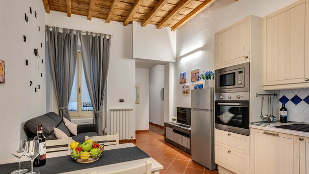 A modern kitchen with white cabinets, a built-in microwave, and an oven. There is a dining area with a table set for two, a bottle of wine, and a fruit basket. - Boboli Guest House Centro Storico Fiorentino (Firenze)